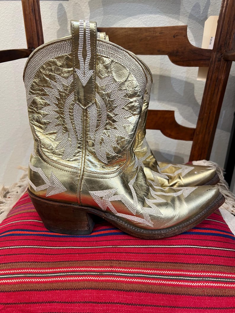 May include: A metallic gold cowboy boot featuring a pointed toe, a stacked wooden heel, and intricate white stitched patterns across the shaft and vamp. The boot has a textured finish and a pull tab at the top of the shaft for easy wear.