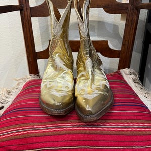 May include: A pair of metallic gold cowboy boots featuring white stitched embroidery patterns on the shaft and vamp. The boots have pointed toes, stacked wooden heels, and pull tabs at the top of the shaft, showcasing a classic western silhouette.