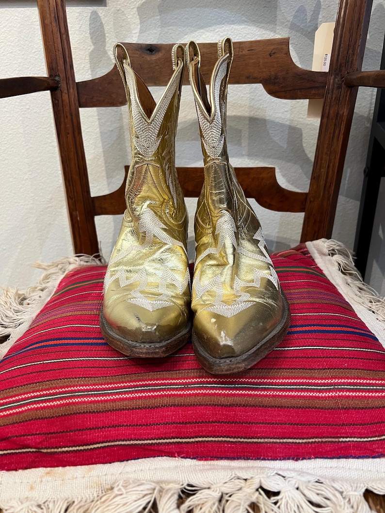May include: A pair of metallic gold cowboy boots featuring white stitched embroidery patterns on the shaft and vamp. The boots have pointed toes, stacked wooden heels, and pull tabs at the top of the shaft, showcasing a classic western silhouette.