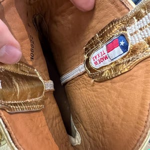 May include: The interior of a boot featuring tan leather lining and metallic gold trim along the top edge. A rectangular fabric label is stitched inside, displaying the Texas state flag and the text "MADE IN TEXAS". The number 108622 is printed.