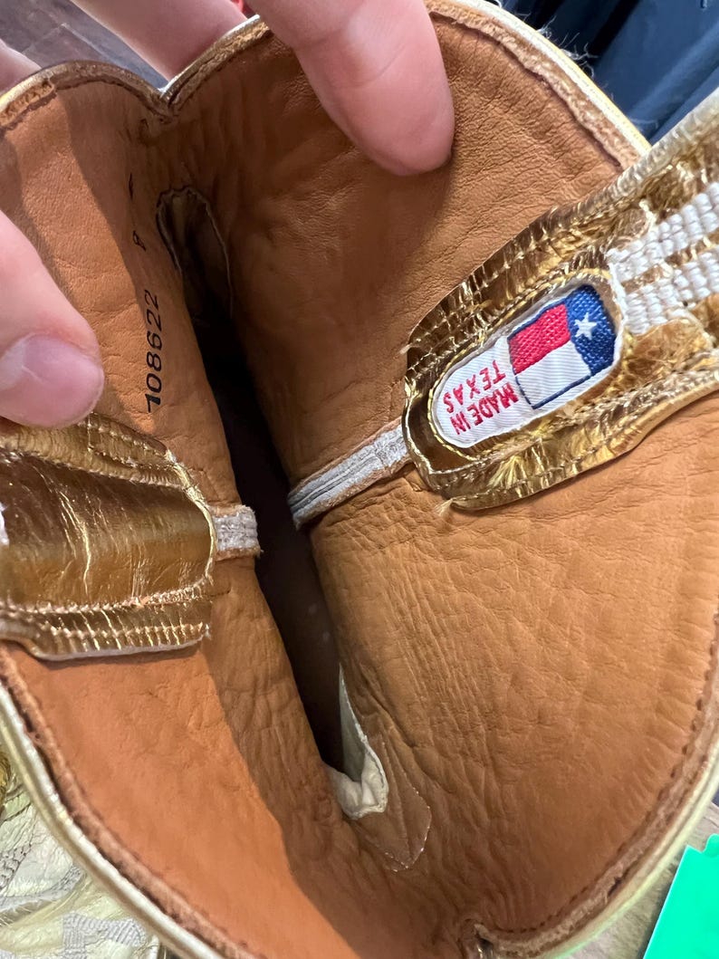 May include: The interior of a boot featuring tan leather lining and metallic gold trim along the top edge. A rectangular fabric label is stitched inside, displaying the Texas state flag and the text "MADE IN TEXAS". The number 108622 is printed.