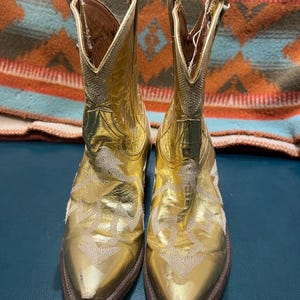 May include: A pair of metallic gold cowboy boots featuring a pointed toe, a mid-calf shaft, and intricate white stitching patterns across the vamp and sides. The boots have a stacked brown heel and a pull tab at the top of each shaft.