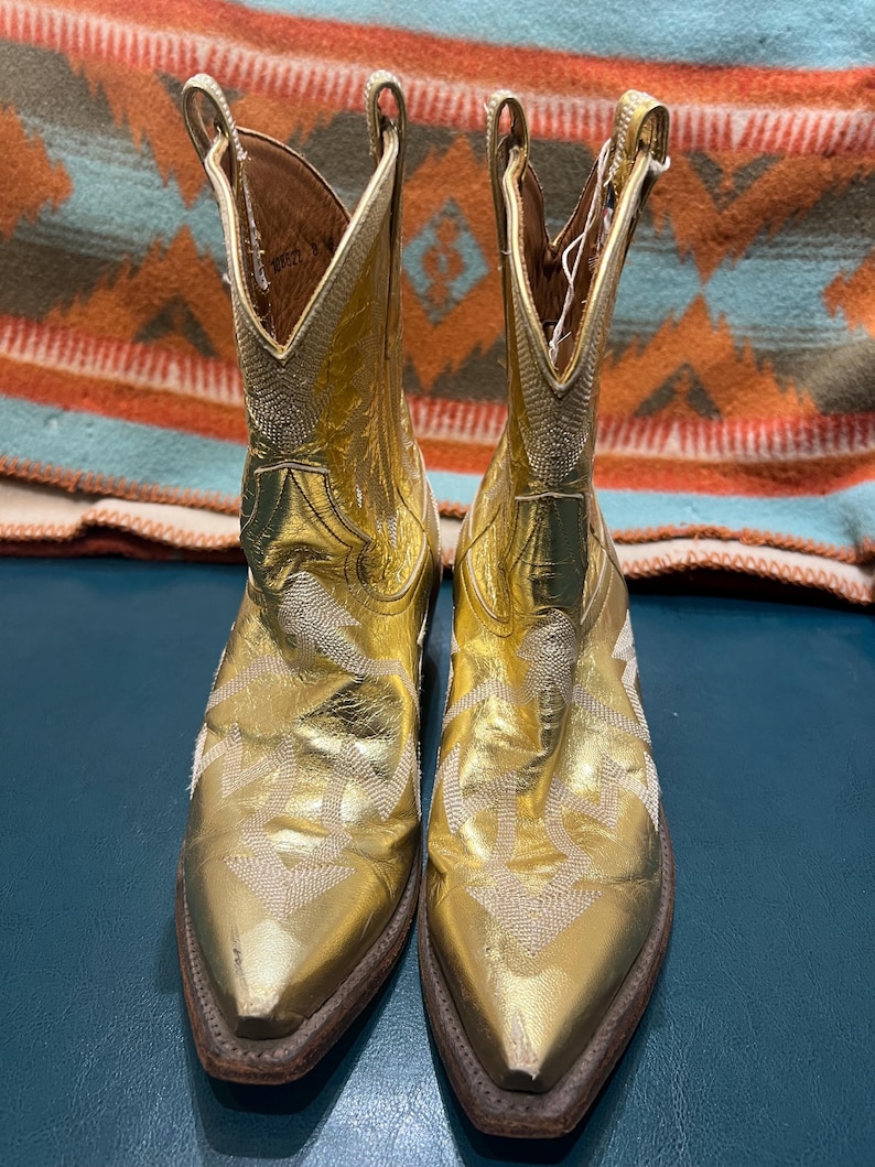 May include: A pair of metallic gold cowboy boots featuring a pointed toe, a mid-calf shaft, and intricate white stitching patterns across the vamp and sides. The boots have a stacked brown heel and a pull tab at the top of each shaft.