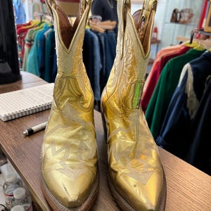May include: A pair of metallic gold cowboy boots featuring a pointed toe, a stacked wooden heel, and intricate white stitched patterns along the shaft and vamp. The boots have pull tabs at the top and a classic western silhouette with a leather finish.