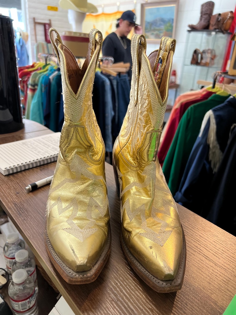 May include: A pair of metallic gold cowboy boots featuring a pointed toe, a stacked wooden heel, and intricate white stitched patterns along the shaft and vamp. The boots have pull tabs at the top and a classic western silhouette with a leather finish.