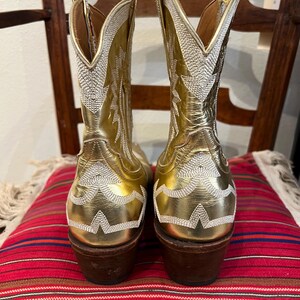 May include: A pair of metallic gold cowboy boots featuring intricate white stitched patterns along the shaft and heel. The boots have a pointed toe, a tall shaft with a V-shaped collar, and a stacked brown wooden heel with a dark rubber base.