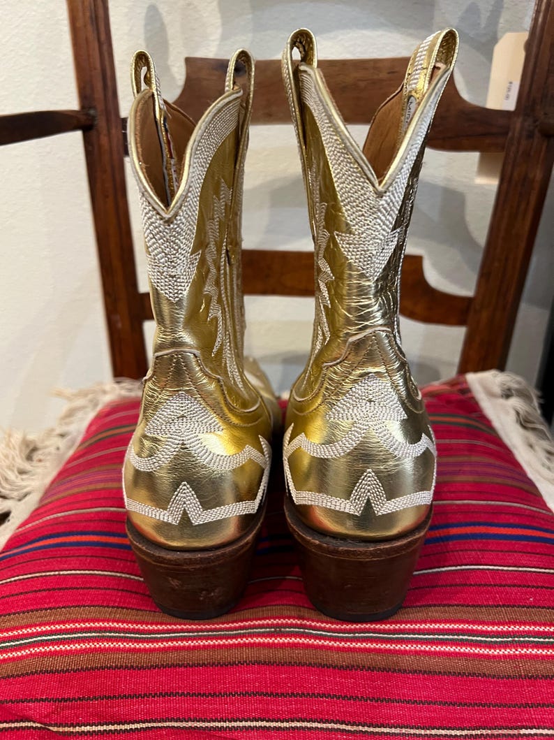 May include: A pair of metallic gold cowboy boots featuring intricate white stitched patterns along the shaft and heel. The boots have a pointed toe, a tall shaft with a V-shaped collar, and a stacked brown wooden heel with a dark rubber base.