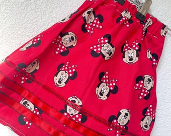 May include: A red skirt featuring a repeating pattern of Minnie Mouse's face with a polka dot bow. The skirt has red ribbon accents and is hanging on a hanger. The skirt is made of a lightweight fabric.