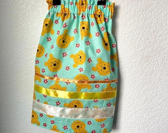 May include: A light blue skirt featuring a repeating pattern of yellow Winnie the Pooh faces and pink flowers. The skirt has gold and yellow ribbon accents, and a white patterned ribbon near the hem. The skirt is hanging on a metal hanger.