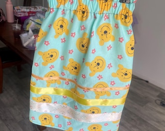 May include: A light blue skirt featuring a repeating pattern of yellow Winnie the Pooh faces and pink flowers. The skirt is adorned with yellow and white ribbons. The skirt is hanging from a metal hanger.