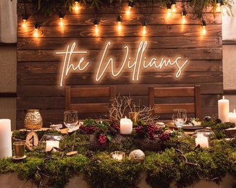 May include: A rustic wedding table setting with a wooden backdrop decorated with string lights and a neon sign that reads "the Williams". The table is adorned with moss, greenery, candles, and red berries.
