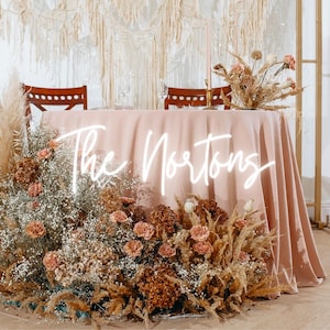 Könnte beinhalten: Eine Hochzeitsempfangstisch-Dekoration mit einer rosafarbenen Tischdecke und einem Neon-Schild mit der Aufschrift "The Nortons". Der Tisch ist mit Trockenblumen und Pampasgras dekoriert.