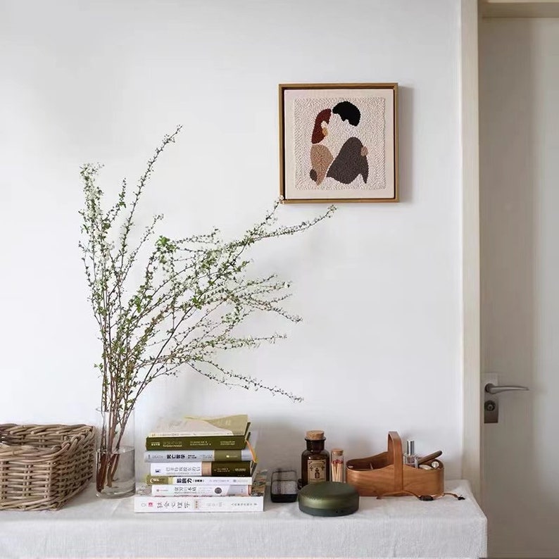 Puede incluir: Una pared blanca con una obra de arte de punto de aguja enmarcada de una pareja abraz&aacute;ndose. Un jarr&oacute;n con una rama de flores blancas se encuentra sobre una mesa con una pila de libros, una caja de madera, una peque&ntilde;a botella y un objeto redondo verde.