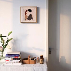 Puede incluir: Una obra de arte enmarcada con un fondo beige y un dise&ntilde;o abstracto colorido de dos figuras en un abrazo rom&aacute;ntico. La obra de arte est&aacute; colgada en una pared blanca sobre una mesa con una pila de libros, una caja de madera y una botella de vidrio.