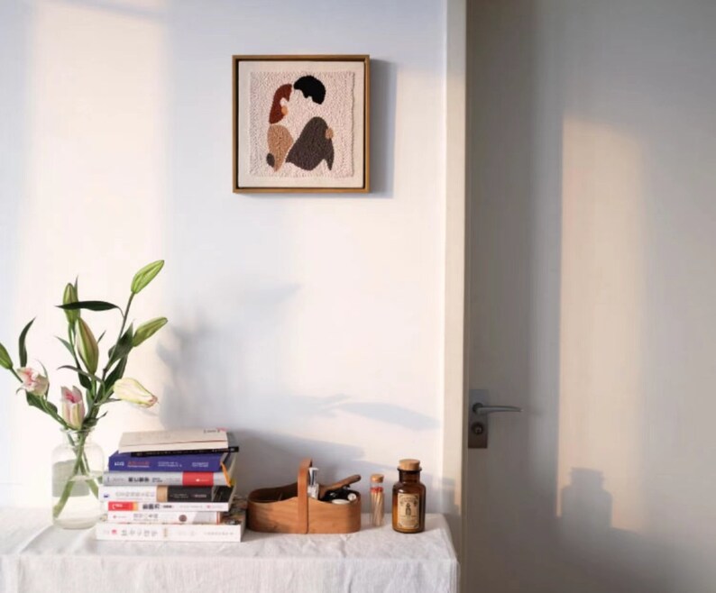 Puede incluir: Una obra de arte enmarcada con un fondo beige y un dise&ntilde;o abstracto colorido de dos figuras en un abrazo rom&aacute;ntico. La obra de arte est&aacute; colgada en una pared blanca sobre una mesa con una pila de libros, una caja de madera y una botella de vidrio.