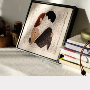 Puede incluir: Una obra de arte de punto de aguja enmarcada que representa dos figuras en formas abstractas, con hilo marr&oacute;n, beige y negro. La obra de arte est&aacute; en un estante blanco con una cesta de madera, botellas y una pila de libros con un altavoz verde en la parte superior.