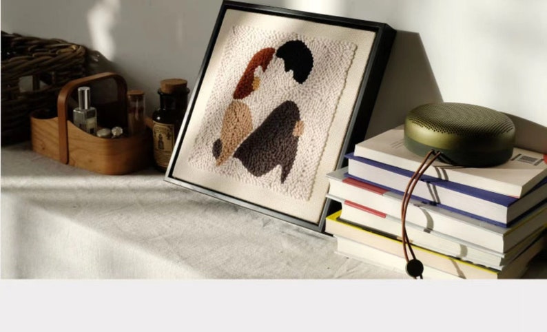 Puede incluir: Una obra de arte de punto de aguja enmarcada que representa dos figuras en formas abstractas, con hilo marr&oacute;n, beige y negro. La obra de arte est&aacute; en un estante blanco con una cesta de madera, botellas y una pila de libros con un altavoz verde en la parte superior.