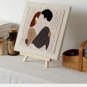 Puede incluir: Un lienzo beige con un dise&ntilde;o de aguja de punz&oacute;n de dos figuras abstractas en marr&oacute;n, beige, gris y negro. El lienzo est&aacute; montado en un peque&ntilde;o caballete de madera.