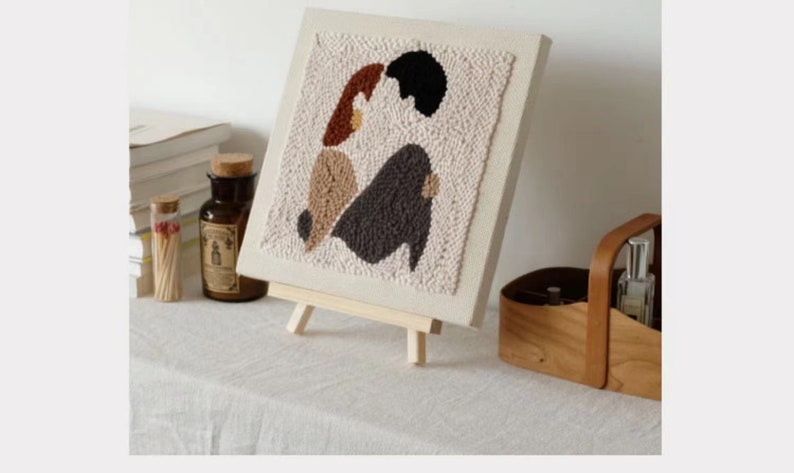 Puede incluir: Un lienzo beige con un dise&ntilde;o de aguja de punz&oacute;n de dos figuras abstractas en marr&oacute;n, beige, gris y negro. El lienzo est&aacute; montado en un peque&ntilde;o caballete de madera.