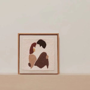 Puede incluir: Una obra de arte enmarcada que representa a una pareja abraz&aacute;ndose, creada con una t&eacute;cnica de aguja de punz&oacute;n. La obra est&aacute; sobre un lienzo blanco con un marco de madera y presenta colores marrones, beige y rojos.
