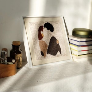 Puede incluir: Una obra de arte de punto de aguja enmarcada que representa a una pareja abraz&aacute;ndose. La obra est&aacute; hecha con varios tonos de hilo marr&oacute;n, beige y blanco. La obra se exhibe en un estante blanco con otros art&iacute;culos decorativos.