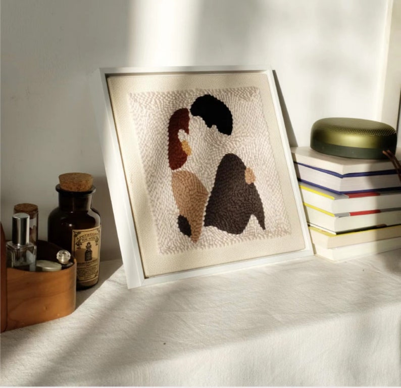 Puede incluir: Una obra de arte de punto de aguja enmarcada que representa a una pareja abraz&aacute;ndose. La obra est&aacute; hecha con varios tonos de hilo marr&oacute;n, beige y blanco. La obra se exhibe en un estante blanco con otros art&iacute;culos decorativos.