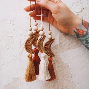Könnte beinhalten: Dekorativer Hängeornament mit gewebten Halbmonden, Holzperlen und Quasten in den Farbtönen Beige, Weiß und Rost. Das Ornament wird an Schnüren gehalten und vor einer strukturierten weißen Wand gehalten.