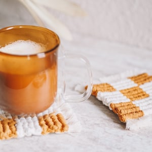 Könnte beinhalten: Eine klare Glas-Tasse, gefüllt mit einem hellbraunen Getränk, steht auf einem weiß-goldenen Makramee-Untersetzer. Ein weiterer Untersetzer ist sichtbar, zusammen mit einer getrockneten Pflanze. Die Untersetzer haben ein Schachbrettmuster.