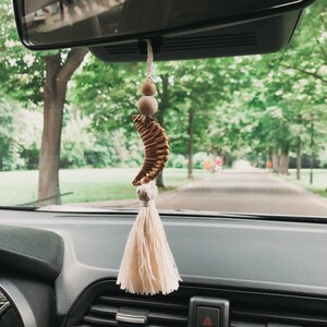 以下が含まれることがあります： バックミラーから吊り下げられた車の芳香剤。木製のビーズ、織り込まれた三日月型の要素、クリーム色のタッセルが特徴です。背景には並木道が写っています。車両用の装飾品です。
