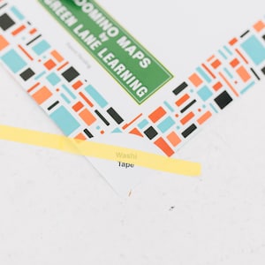 May include: A white card with a geometric border of orange, light blue, and black rectangles. The text reads DOMINO MAPS BY GREEN LANE LEARNING. A thin strip of yellow washi tape is placed across the corner of the card, partially obscuring the text.