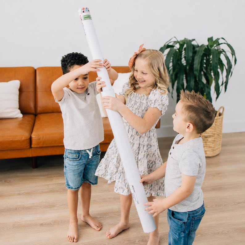 May include: A long, white cylindrical cardboard shipping tube with a small label at the top. The tube is held vertically by three children who are standing together in a room with light wood flooring and a tan leather sofa in the background.