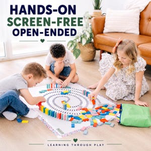 May include: A set of colorful wooden dominoes arranged on a white play mat with a spiral pattern. The mat features a floral border and is accompanied by a green storage bag. Text on the image reads: HANDS-ON SCREEN-FREE OPEN-ENDED and LEARNING THROUGH PLAY.
