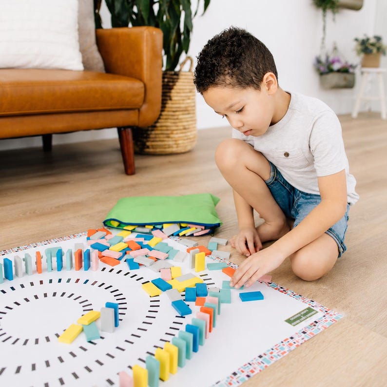 May include: A white play mat featuring a circular black dotted pattern for arranging colorful wooden dominoes. The set includes a green storage bag and numerous rectangular dominoes in shades of blue, yellow, pink, and gray for building.