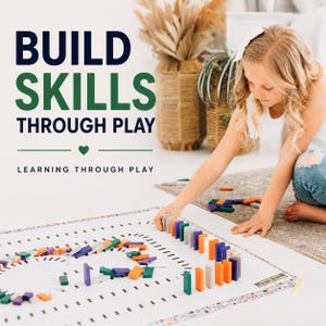 May include: A white play mat with black dashed lines and a colorful set of rectangular dominoes arranged in a track. The text reads BUILD SKILLS THROUGH PLAY and LEARNING THROUGH PLAY with a small green heart icon between the two phrases.