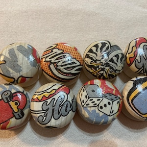 May include: Nine round wooden knobs with colourful designs. The designs include a skateboard, hot dog, dice, a television, a pair of sneakers, and the words "New York", "Florida", and "Crazy".