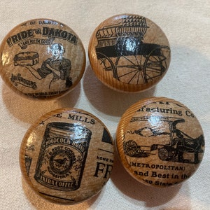 May include: Four wooden knobs with vintage advertisements printed on them. The knobs feature images of a horse-drawn carriage, a coffee can, a steam engine, and a woman holding a loaf of bread. The text on the knobs includes "Pride of Dakota", "Take Me In Good", "Woodcock Brand", "E. Mills", "Home Products", "Manufacturing Co", "Metropolitan", and "Best in the 20 States".