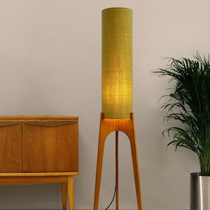 May include: A tall, mid-century modern floor lamp with a cylindrical, olive green fabric shade. The lamp has a wooden tripod base and a black power cord. The lamp is next to a wooden cabinet and a potted plant.