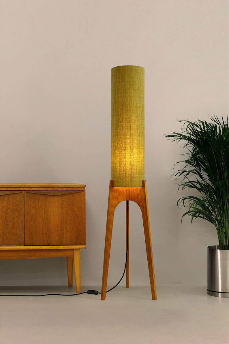 May include: A tall, mid-century modern floor lamp with a cylindrical, olive green fabric shade. The lamp has a wooden tripod base and a black power cord. The lamp is next to a wooden cabinet and a potted plant.
