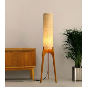May include: A mid-century modern floor lamp with a cylindrical, beige fabric shade. The lamp has a wooden tripod base and a warm, inviting glow. A wooden cabinet and a potted plant are in the background.