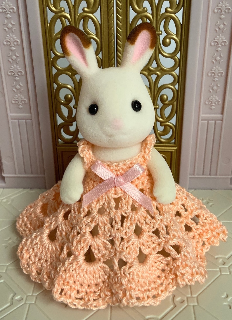 May include: A white rabbit doll wearing a peach crocheted dress with a pink ribbon.