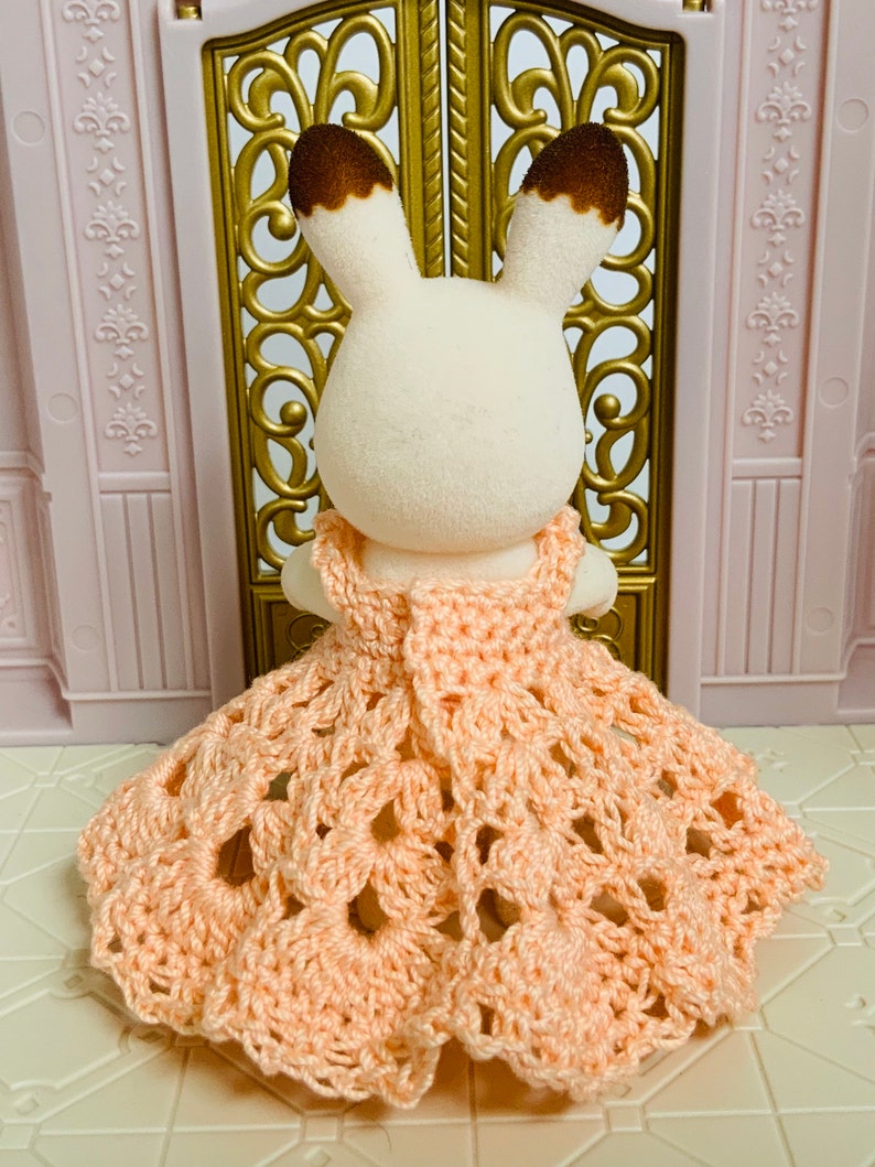 May include: A small, white rabbit wearing a peach-colored crocheted dress with a scalloped hem.