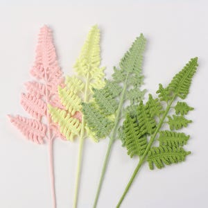 May include: Four crochet fern stems in shades of pink, yellow, and green. The fern leaves have a detailed, textured appearance. The stems are a solid color, and the leaves are a lighter shade of the same color. The ferns are arranged against a white background.