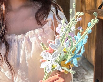 Puede incluir: Un ramo de flores de ganchillo hechas a mano en varios colores, incluyendo blanco, azul, amarillo, rosa y morado. Las flores son sostenidas en las manos de una persona, con una blusa blanca en el fondo.