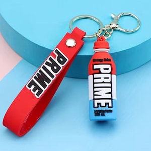 Könnte beinhalten: Rot-blauer Prime Energy Drink Schlüsselanhänger mit einem roten Band, auf dem "PRIME" in schwarzen Buchstaben steht. Der Schlüsselanhänger hat einen silbernen Schlüsselring.