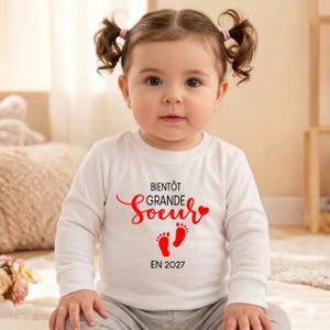 Peut inclure: Un bébé porte un t-shirt blanc à manches longues avec l'inscription "BIENTÔT GRANDE Soeur" en rouge, ainsi que des empreintes de pieds rouges et l'année "EN 2027". Le bébé a les cheveux bruns en couettes.