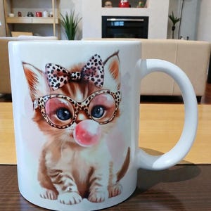 May include: White ceramic mug featuring a cartoon kitten with blue eyes, wearing glasses and a bow. The kitten is blowing a pink bubble. The mug has a white handle and is likely used for beverages.