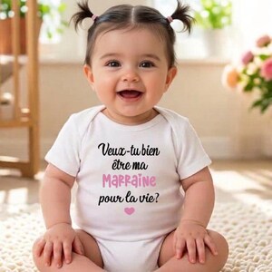 May include: A smiling baby wearing a white onesie with the French text "Veux-tu bien &ecirc;tre ma Marraine pour la vie?" in black and pink, with a pink heart. The baby has dark hair in pigtails.