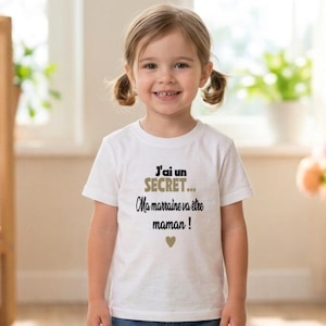 Peut inclure: T-shirt blanc pour enfant avec l'inscription "J'ai un SECRET... Ma marraine va être maman !" en noir et or, et un petit cœur. La chemise est portée par une jeune fille blonde.