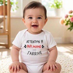 May include: A baby wearing a white onesie with the French text "ATTENTION!!! PAPI, MAMIE... J'ARRIVE" in black and red. The baby is smiling. The baby is sitting on a cream-colored surface.