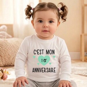 May include: A baby wearing a white long-sleeved shirt with the French text "C'EST MON 1er ANNIVERSAIRE" in black. A turquoise heart and small turquoise hearts are also on the shirt. The baby has brown hair and is looking at the camera.